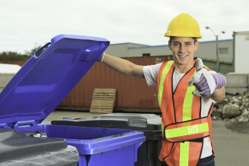 A young male waste management worker wearing a yellow safety helmet, high-visibility orange and yellow vest, and purple gloves is standing outdoors, smiling, next to a large blue recycling bin with its lid open. Behind him, there are industrial buildings and a clear sky, indicating a commercial or industrial area, possibly within or near Bow. The worker appears to be preparing for rubbish collection service, with his right hand resting on the bin and his left hand giving a thumbs-up gesture. The scene reflects a professional approach to waste removal, with the worker ready to assist with rubbish clearance tasks typical of commercial waste services provided by companies like Commercial Waste Bow in the local Bow area. The environment suggests an organized waste collection operation, emphasizing safety and efficiency in handling general rubbish or recyclable materials in a business context.