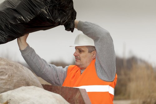 A male waste collection worker wearing a white safety helmet and an orange high-visibility vest is lifting a large black rubbish bag with both hands, positioning it above a pile of various rubbish materials. The rubbish pile includes large, beige-colored sacks and smaller debris, situated outdoors on a paved or concrete surface. In the background, there is a blurred natural landscape with some trees or shrubs, indicating an open environment typical for waste collection services around Bow or the surrounding East London area. The scene is lit with natural daylight, suggesting it is daytime, and the worker appears focused on the task of rubbish removal. This image visually supports the services offered by Commercial Waste Bow, specializing in professional rubbish collection and waste management for local businesses in the postcode area.