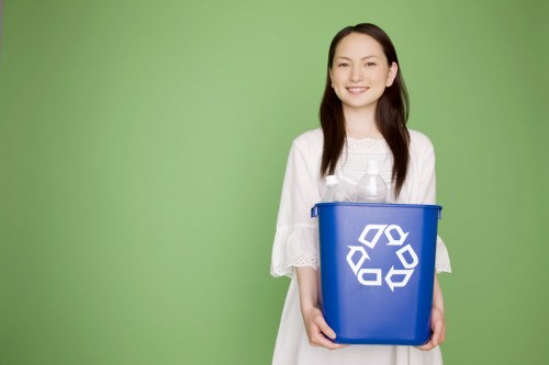 A young woman with long dark hair and a friendly expression stands against a plain green background, wearing a white dress with lace details on the sleeves. She holds a blue recycling bin with a white recycling symbol printed on the front in both hands, positioned in front of her torso. The bin is made of plastic, with a slightly textured surface, and appears to be empty or containing recyclable waste. Her stance suggests she is presenting or demonstrating disposal of recyclable rubbish. The setting is simple and uncluttered, typical of a practitioner or promotional image related to waste management services in Bow, within the London area. The image conveys themes of environmental responsibility and proper waste segregation, aligning with services provided by Commercial Waste Bow for commercial rubbish collection and recycling efforts.
