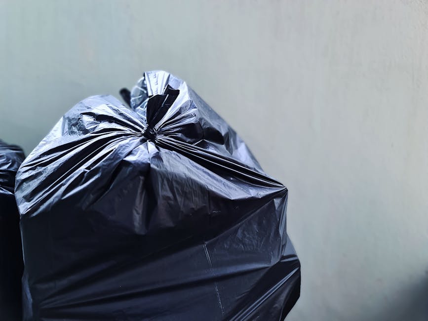 The image displays several black plastic rubbish bags, tightly knotted at the top, stacked against a neutral-colored wall. The bags are made of glossy, crinkled plastic material, reflecting light and 