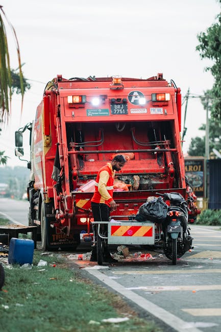 A street scene showing a waste collection vehicle with a red and yellow color scheme, parked on the side of the road. In front of the vehicle, a worker wearing high-visibility orange and yellow clothi